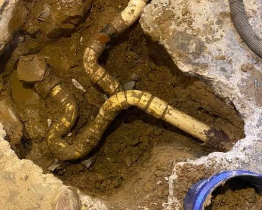 Prodigy Sewer and Drain working on underground pipes for repairs in a dirt-filled area under a concrete floor.